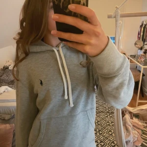 Grå hoodie från Polo Ralph Lauren - Snygg ljusgrå hoodie från Polo Ralph Lauren med klassisk broderad logga på bröstet. Tröjan har huva med vita snören och en stor magficka. Tillverkad i mjuk bomull, perfekt för chill dagar. Passformen är normal och ärmarna är långa. Storlek är S men skulle säga att den passar M också. Vid frågor det självklart bara att fråga!😁🤙🏻