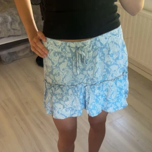 Blå kortkjol med volang och knyt - Supersöt ljusblå kortkjol med vitt paisleymönster och volangkant. Kjolen har knytband i midjan och har insydda shorts. Har använt kjolen en sommar men har hållt den i ett bra skick💗