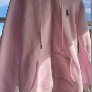 Ljusrosa hoodie! - Supermjuk ljusrosa hoodie med dragkedja, huva och snörning. Logga på bröstet och stora fickor framtill. Tillverkad i bomullsmix med fleeceinsida för extra komfort. Perfekt för chill dagar eller när du vill vara extra cozy.