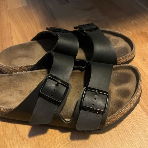 Svarta sandaler från Birkenstock - stl 41 - Klassiska svarta sandaler från Birkenstock med två breda remmar i skinn och justerbara svarta spännen. Fotbädden är formad för extra komfort och sulan är i svart gummi med mönster. Sulorna något slitna men inget som stör. Perfekta för sommaren och riktigt sköna att gå i.
