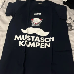 Mörkblå Mustaschkampen t-shirt XS - Snygg mörkblå t-shirt från B&C Collection i storlek XS med stort vitt tryck 'Mustaschkampen', mustaschmotiv och Linköping Hockey Club-logga. Klassisk passform och korta ärmar, perfekt för dig som vill sticka ut med stil och visa ditt stöd.