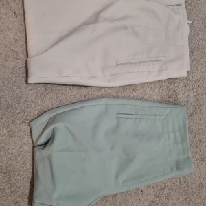 Ljusgröna och vita kostymbyxor - Två par stilrena kostymbyxor i ljusgrönt och vitt. Byxorna har hög midja, raka ben och pressveck framtill. Snygga fickor i sidorna och minimalistisk design. Perfekta för dig som gillar en clean och modern look.