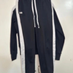 Svartvitt tracksuit från Under Armour - Svarta mjukisbyxor från Under Armour med vita sidoränder och dragsko i midjan. Byxorna har resår vid benslut och en sportig look, perfekta för chill eller träning. Materialet är mjukt och stretchigt för extra komfort.