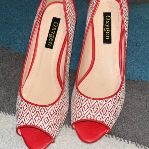 Rödvita pumps från Oxygen med mönster - Snygga pumps från Oxygen i rödvitt med geometriskt mönster. Skorna har öppen tå, låg klack och röda detaljer längs kanterna. Perfekta för dig som vill sticka ut med en unik och färgglad stil.