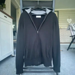 Svart hoodie från Pier One med dragkedja - Svart hoodie från Pier One med hel dragkedja framtill och två fickor. Insidan av huvan är ljusgrå och ribbad, vilket ger en snygg kontrast. Perfekt för dig som gillar en enkel och stilren look. Långärmad och med klassisk passform.