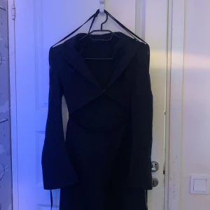 Svart jumpsuit från PrettyLittleThing - Snygg svart jumpsuit från PrettyLittleThing med långärmad design och en unik öppen fram detalj. Perfekt för en elegant look och festlig look. Det finns ’pads’ vid axlarna. Storlek 38 men är inte stretchy. Klänningen har en figurnära passform och är tillverkad i ett mjukt material.