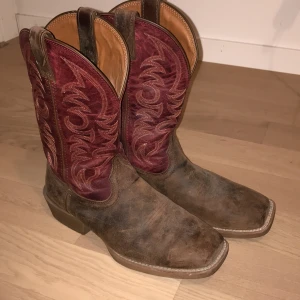Bruna och vinröda cowboy boots - Snygga cowboy boots i brunt skinn med vinröda broderade detaljer. De har en fyrkantig tå och en robust klack. Perfekta för en stilren look med en touch av western.