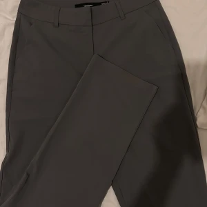 Grå kostymbyxor från Vero Moda - Snygga grå kostymbyxor från Vero Moda med en klassisk design. Byxorna har en rak passform och är perfekta för en stilren look. De har hällor för bälte och en diskret dragkedja framtill.