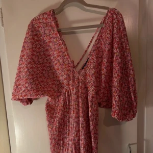 Blommig klänning med puffärmar - Charmig blommig klänning i rosa och vita toner med puffiga ärmar och v-ringning. Perfekt för en somrig look med sitt lätta och luftiga material. Klänningen har en omlott-design som ger en fin siluett.