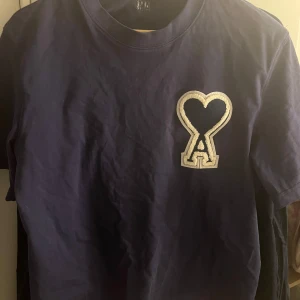 Blå t-shirt från Ami Paris  - Säljer en snygg blå t-shirt från Ami med ett coolt hjärtmönster och bokstaven A på bröstet. Perfekt för en avslappnad stil. T-shirten har korta ärmar och en klassisk rund halsringning. Lite mindre i storleken. 