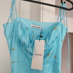 Blå Adoore klänning i storlek 36 - Sommarklänning från Adoore men härligt blått mönster med gula små blommor på. Materialet känns siden-aktigt. Storlek 36. Band att knyta vid axlar och byst för att passa just din kropp. Finns en dragsko på sidan också som gör att man kan välja längd på klänningen efter eget tycke. Helt ny med lapp kvar.