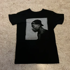 Svart T-shirt med 2Pac-tryck - Säljer en svart T-shirt med ett stort tryck av 2Pac i profil på framsidan. T-shirten är kortärmad och har en klassisk rund hals. Perfekt för fans av hiphop och streetwear.