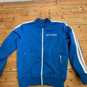 Blå track jacket från Palm Angels - Säljer en snygg blå track jacket från Palm Angels med vita detaljer. Jackan har dragkedja framtill och vita ränder längs ärmarna.  Endast använd ett fåtal gånger. ÄKTA!  // Ian 