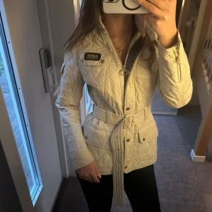 Beige quiltad jacka från Barbour - Snygg beige quiltad jacka från Barbour International med dragkedja och bälte i midjan. Jackan har långa ärmar och stilrena detaljer med knappar. Perfekt för en chic look!