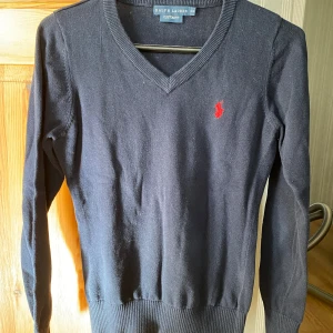 Svart tröja från Ralph Lauren - Säljer en stilren svart tröja från Ralph Lauren med v-ringning och röd broderad logga på bröstet. Tröjan har långa ärmar och ribbade muddar vid ärmslut och nederkant. Perfekt för en klassisk look.