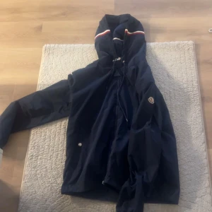Moncler windbreaker  - Säljer en mörkblå vindjacka med huva och dragkedja. Jackan har vita och röda ränder på huvan och dragsko vid halsen. Perfekt för blåsiga dagar!. Storlek 4 så L/M