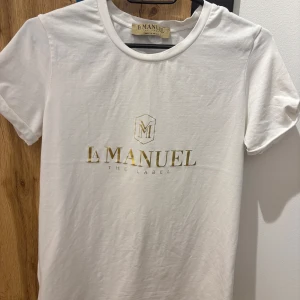Vit t-shirt från La Manuel - Säljer en stilren vit t-shirt från La Manuel med guldig logga på framsidan. Perfekt för en enkel och snygg look. Tillverkad i Polen.