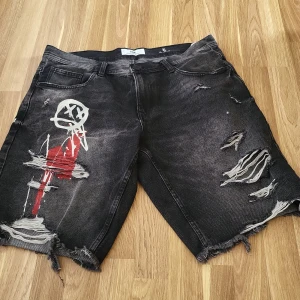 Svarta jeansshorts från FSBN - Säljer ett par svarta jeansshorts från FSBN med slitna detaljer och tryck. Shortsen har en cool, distressed look med fransar och konstnärligt mönster i rött och vitt. Perfekta för en avslappnad stil.
