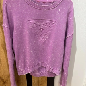Lila tröja från Guess - Säljer en cool lila tröja från Guess med deras ikoniska logga på framsidan. Tröjan har en avslappnad passform och är perfekt för en casual look. Den är långärmad och har en lätt tvättad effekt som ger en vintage känsla.