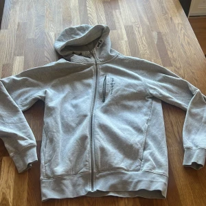 Grå zip hoodie från Sail Racing - Säljer en stilren grå hoodie från Sail Racing med dragkedja och huva. Perfekt för en avslappnad look. Den har en praktisk bröstficka med dragkedja och ribbade muddar vid ärmslut och nederkant.
