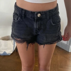 Svarta jeansshorts från Zara - Snygga svarta jeansshorts från Zara med slitna detaljer. De har en klassisk femficksdesign och knappgylf. Säljs inte längre och är väldigt lågmidjade😍