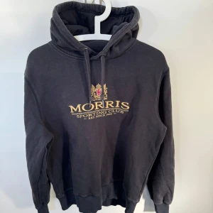 Mörkblå hoodie från Morris - Säljer en stilren hoodie från Morris med broderad logotyp på bröstet. Tröjan har en klassisk design med justerbar huva och ribbade muddar vid ärmslut och nederkant. Perfekt för en avslappnad look. Org pris 1500