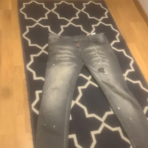 Grå jeans med slitningar - Säljer ett par grå jeans med slitningar och färgstänk. De har en klassisk femficksdesign och en patch på baksidan. Perfekta för en avslappnad stil.