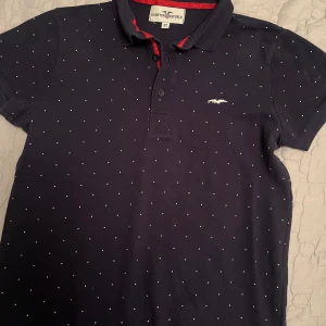 Mörkblå pikétröja från Hampton Republic - Snygg mörkblå pikétröja från Hampton Republic med små vita prickar och en broderad logga på bröstet. Tröjan har en klassisk krage med röda detaljer och knappar framtill. Perfekt för en stilren look!