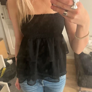 Svart axelbandslös topp - Snygg svart axelbandslös topp med volanger. Perfekt för en kväll ute eller en speciell tillställning. Den har en smockad överdel som ger en bra passform med volanger nedantill, kan också användas som kjol. Endast använd 1 gång🩷