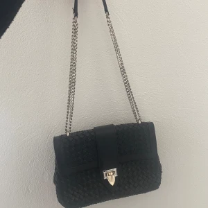 Svart axelväska från Decadent - Snygg svart axelväska från Decadent med flätat mönster och en elegant kedjerem. Väskan har en stilren låsdetalj i silver/guld och är perfekt för att ge en touch av lyx till din outfit.