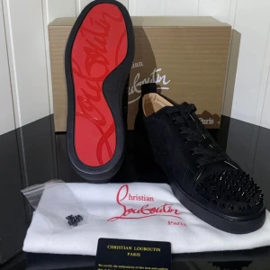 Svarta sneakers med nitar från Christian Louboutin - Snygga svarta sneakers från Christian Louboutin med ikoniska röda sulor och nitar på ovansidan. Skorna har snörning och en rund tå, vilket ger en edgy och modern look. Perfekta för att sticka ut i mängden. Använd dom inte sår ofta 