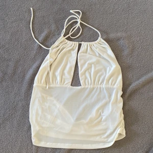Vit ärmlös halterneck-topp - Säljer en stilren vit halterneck-topp med knytband i nacken. Toppen har en öppen rygg och en liten öppning framtill som ger en snygg detalj. Perfekt för varma sommardagar eller en kväll ute. Passar bra till både jeans och kjol. Stl xl, köpt på Gina. Finns en fläck som jag inte vet om den går bort i tvätt (se bild), inget jag tänkt på men därav lågt pris.