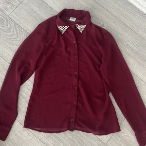Vinröd blus från Vero Moda - Snygg vinröd blus från Vero Moda med långa ärmar och pärldetaljer på kragen. Perfekt för att ge en elegant touch till din outfit. Blusen har knappar framtill och är tillverkad i ett lätt och bekvämt material.