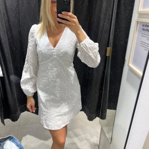 Vit klänning med puffärmar - Snygg vit klänning med v-ringning och puffiga långärmar. Klänningen har ett subtilt blommönster och en kort längd som ger en fräsch look. Perfekt för sommarens och även studenten🥰
