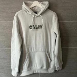 Vit hoodie med texten 'CALM' - Säljer en stilren vit hoodie med texten 'CALM' broderad på bröstet. Den har en klassisk känguruficka och justerbar huva med dragsko. Perfekt för en avslappnad look.