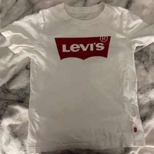 Vit t-shirt från Levi's - Säljer en klassisk vit t-shirt från Levi's med det klassiska röda logotyptrycket på bröstet. T-shirten har en normal passform och är kortärmad. Den har en fläck, som knappt syns. Men ifall man kollar noga så ser man den. T-shirten är använd några gånger💕