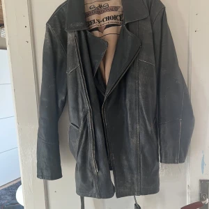 Svart skinnjacka från Rebel's Choice - Säljer en svart skinnjacka från Rebel's Choice med dragkedja och bälte i midjan. Jackan har en klassisk design med långa ärmar och en stilren look. Perfekt för en cool och tidlös stil.