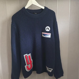 Mörkblå stickad tröja från J.Lindeberg - Säljer en snygg mörkblå stickad tröja från J.Lindeberg med coola patches på framsidan. Tröjan har en ribbad design och långa ärmar, perfekt för kyligare dagar. Passar bra för en stilren och avslappnad look.