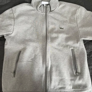 Grå tröja från Lacoste - Säljer en stilren grå tröja från Lacoste med dragkedja och det klassiska krokodilmärket på bröstet. Jackan har långa ärmar och praktiska fickor med dragkedjor. Perfekt för en casual look.