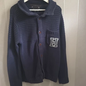 Mörkblå stickad kofta från Hilfiger  - Säljer en snygg mörkblå stickad kofta från Hilfiger  med knappar framtill och en cool H-applikation på bröstet. Koftan har en ribbad krage och långa ärmar, perfekt för kyliga dagar. Passar både till jeans och chinos!