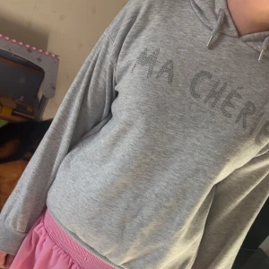 Grå hoodie med text - Säljer en grå hoodie med texten 'MA CHÉRIE' i glittriga stenar på framsidan. Tröjan har en bekväm passform med långa ärmar och en justerbar huva. Perfekt för en avslappnad stil.