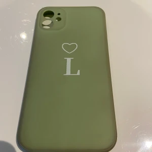 Grönt mobilskal med hjärta och bokstav - Snyggt grönt mobilskal med ett stilrent hjärta och bokstaven 'L' i vitt. Perfekt för att ge din mobil en personlig touch. Skalet är designat för att skydda baksidan av din telefon och har utskärningar för kamera och knappar. iPhone 11 