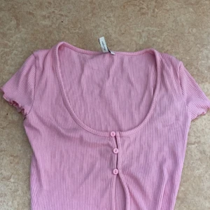 Rosa ribbad topp från Nelly - Säljer en söt rosa ribbad topp från Nelly med korta ärmar och knappar framtill. Perfekt för en avslappnad stil. 💖
