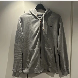 Grå hoodie från Morris - Säljer en stilren grå hoodie från Morris med dragkedja och justerbar huva. Tröjan har långa ärmar och praktiska fickor framtill. Perfekt för en avslappnad look. Pris kan diskuteras vid snabb och simpel affär