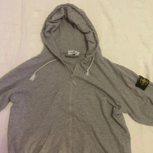 Grå hoodie från Stone Island - Säljer en stilren grå hoodie från Stone Island med dragkedja och justerbar huva. Tröjan har en ikonisk logga på ärmen och är perfekt för en avslappnad look. Passar bra till både jeans och joggers. Kan gå ner i pris vid snabb affär.