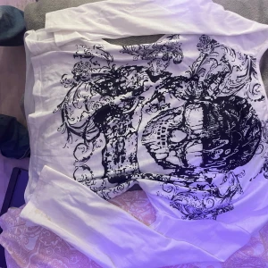 Vit tröja y2k - Säljer en vit långärmad tröja med ett stort svart dödskallemönster på framsidan. Tröjan har en unik design med detaljerade blommor och ornament runt dödskallen. Perfekt för dig som gillar en edgy stil.