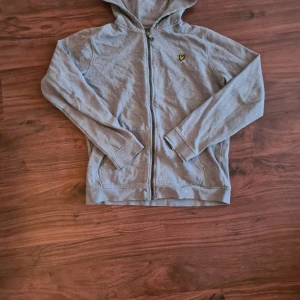 Grå tröja från Lyle & Scott - Säljer en grå tröja från Lyle & Scott med dragkedja och broderad logga på bröstet. Sliten längst ut i ena ärmen, men lika snygg och mysig ändå!