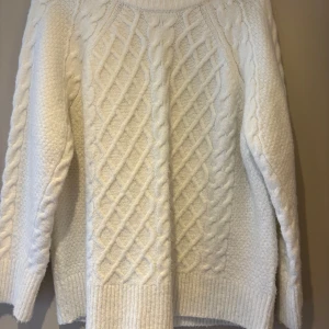 Vit stickad tröja från 157 - Mysig vit stickad tröja från 157 med ett klassiskt flätmönster. Perfekt för kyliga dagar med sin varma och bekväma design. Tröjan har långa ärmar och ribbade kanter för extra komfort.