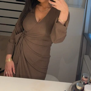 Brun omlottklänning från NA-KD - Snygg brun omlottklänning från NA-KD i ribbat material. Klänningen har långa ärmar och en knytning i midjan som ger en smickrande passform. Perfekt för en stilren look. Använd en gång. 