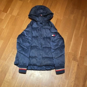 Blå dunjacka från Tommy Hilfiger - Säljer en snygg blå dunjacka från Tommy Hilfiger. Jackan har en huva och långa ärmar med ribbade muddar i rött och vitt. Den är dekorerad med Tommy Hilfigers logotyp på ärmen och bröstet. Perfekt för kyliga dagar!